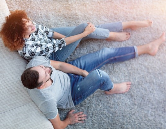 : Couple leaning up against a couch, sitting on carpet flooring. 